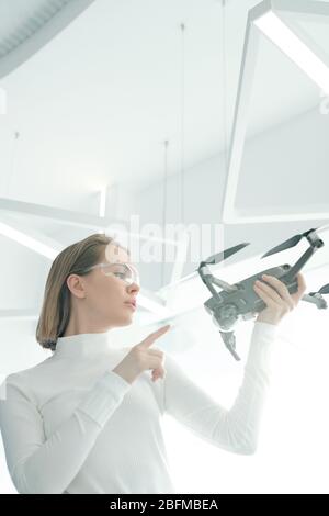 Junge Frau in smarter Brille hält Drohne vor sich und lernt, wie es im futuristischen Labor beginnt und funktioniert Stockfoto