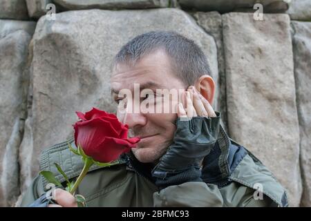 Ein Typ auf der Straße in einer Herbstjacke und Lederhandschuhen sieht kokett aus und schnüffelt eine rote Rosenblume Stockfoto