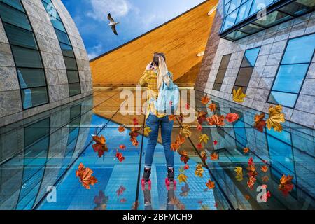 Perspektive. Ein konzeptuelles Foto einer weiblichen Rollerin, die ein Bürogebäude hochfährt, während sie eine Möwe fotografiert, mit hinterhängenden Blättern Stockfoto