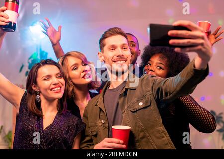 Fröhlicher junger Mann mit Smartphone und drei fröhliche interkulturelle Mädchen machen Selfie und jubeln mit Getränken zu Hause Party Stockfoto