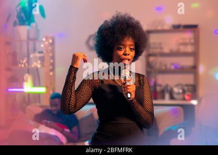 Junge elegante Frau in eleganten schwarzen Kleid singen Karaoke im Mikrofon auf dem Hintergrund von Freunden auf der Couch zu Hause Party Stockfoto