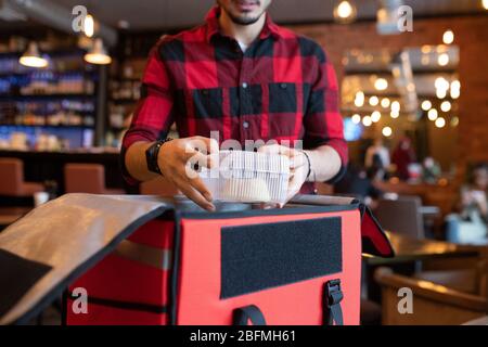 Junge Kurier Putting Kunststoff-Container mit gekochtem Essen in großen offenen Beutel beim Sammeln von Online-Bestellungen von Kunden vor der Auslieferung Stockfoto