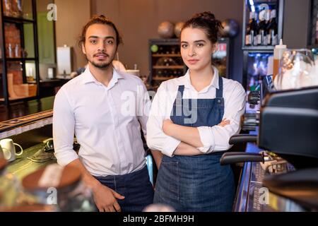 Zwei junge Kollegen in weißen Hemden und Denim-Schürzen stehen am Arbeitsplatz neben der Kaffeemaschine im Café und schauen Sie an Stockfoto
