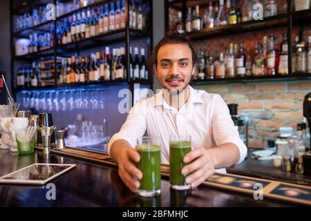 Fröhlicher junger Kellner oder Barkeeper in weißem Hemd, der Ihnen zwei Gläser frisches Gemüse Smoothie mit Flaschen Alkohol auf Hintergrund übergeht Stockfoto