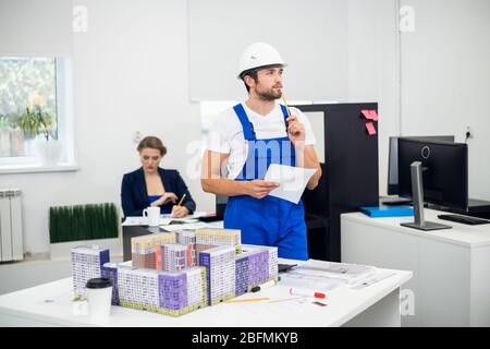 Ein nachdenklicher junger Bauarbeiter im Büro mit einem Baumodell neben ihm. Stockfoto