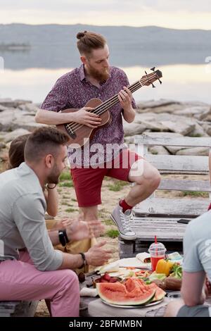 Junge kaukasische bärtige Mann spielt die Gitarre, während mit Freunden aus der Stadt in der Nähe von See an schönen Sommerabend hängen Stockfoto