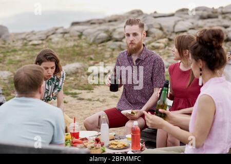 Gruppe von jungen Menschen verbringen Sommerwochenende im Freien, sitzen um den Tisch, essen, Bier trinken und teilen die neuesten Nachrichten Stockfoto
