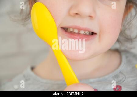 Nettes kleines Mädchen mit holdinging gelben Plastiklöffel in der Hand, lächelt und zeigt Milchzähne. Stockfoto