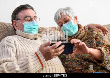 Coronavirus. Bleiben Sie zu Hause, Lifestyle.Fröhliche ältere Paar sitzt auf einem Sofa auf einer Quarantäne zu Hause, einen Videoanruf mit dem Smartphone. Ältere Paar tragen Schutzmasken . Stockfoto