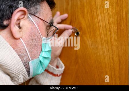 Covid-19-Konzept. Bleiben Sie zu Hause. Selbstisolierung zur Verhinderung der Coronavirus-Pandemie. Senior Mann in schützender grüner Maske schaut durch das Guckloch. Stockfoto