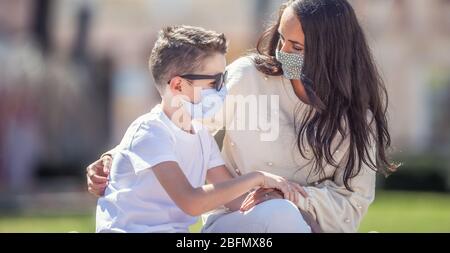 Alleinerziehende Mutter schaut besorgt auf ihren Sohn in Sonnenbrille draußen, beide tragen Gesichtsmasken. Stockfoto