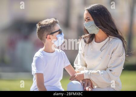 Mutter und Sohn, die Gesichtsmasken tragen, schauen sich gegenseitig an, die draußen sitzen. Stockfoto