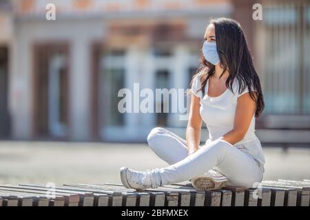 Gut aussehende Mädchen sitzt allein auf der Bank im Stadtzentrum mit einer Gesichtsmaske während Covid-19 Pandemie. Stockfoto