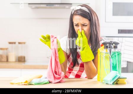 Frau, die ihre Nase mit den Fingern auf stinkenden Lappen in der anderen Hand hält, sich auf den Küchentischtisch lehnt, mit Putzzeug um sie herum. Stockfoto