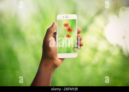 Frau Hand hält Smartphone mit kleinen Blumen auf dem Bildschirm vor unscharfen grünen Hintergrund Stockfoto