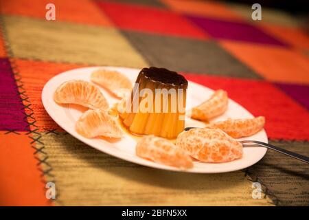 Vanille- und Karamellpudding umgeben von Mandarinensegmenten in einem weißen Gericht auf mehrfarbigem Hintergrund Stockfoto