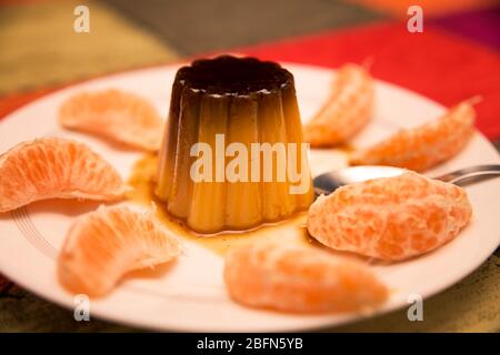 Vanille- und Karamellpudding umgeben von Mandarinensegmenten in einem weißen Gericht auf mehrfarbigem Hintergrund Stockfoto