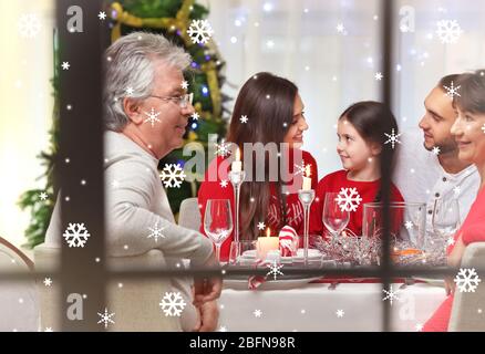 Blick durch das Fenster auf glückliche Familie während des traditionellen Weihnachtsessen. Verschneite Optik. Konzept für Weihnachtsfeiern. Stockfoto