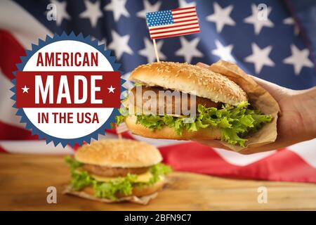 Etikett mit Text AMERICAN MADE IN the USA und männliche Hand mit leckeren Burger auf Flaggenhintergrund Stockfoto