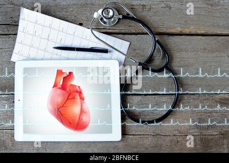 Tablet mit Stethoskop auf Holzhintergrund. Herz auf dem Bildschirm. Medizin und modernes Technologiekonzept. Stockfoto