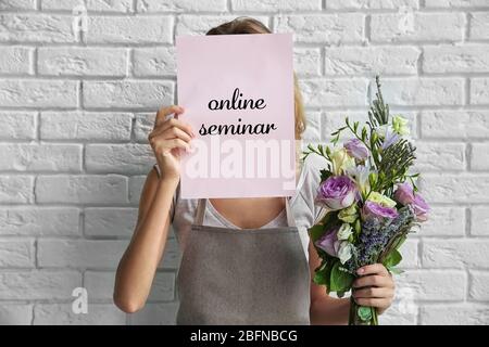 Frau hält Papier mit Text ONLINE-SEMINAR und Strauß auf Backstein Wand Hintergrund. Floristin und Blumendesign Tutorial Konzept. Stockfoto