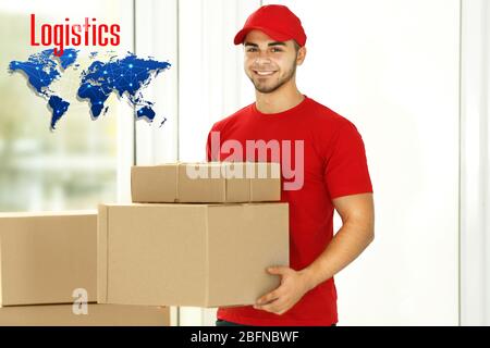 Liefermann in Uniform mit Pappkartons im Lager. Wort LOGISTIK und Weltkarte im Hintergrund. Großhandel und Logistik Konzept. Stockfoto