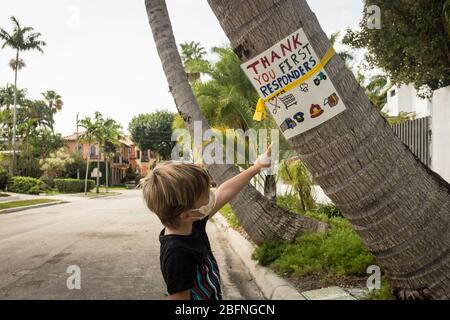 Ein kleiner Junge, der eine Maske trägt, zeigt auf ein Schild auf einer Palme, das sich an die Ersthelfer während der COVID-19-Sperre in Miami, Florida, USA, bedankt. Stockfoto