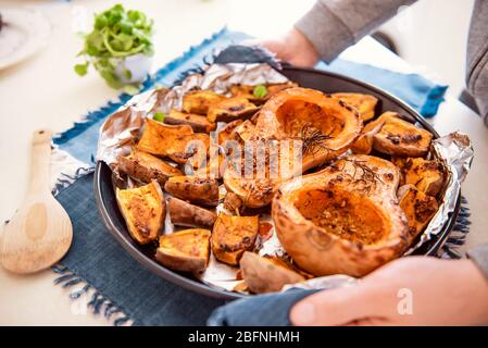 Männliche Hände setzen Backblech mit gerösteten gegrillten orange Kürbis Butternut Kürbis Halbtöne und Süßkartoffel Dias mit Kräutern auf blaue Serviette. Vegetari Stockfoto