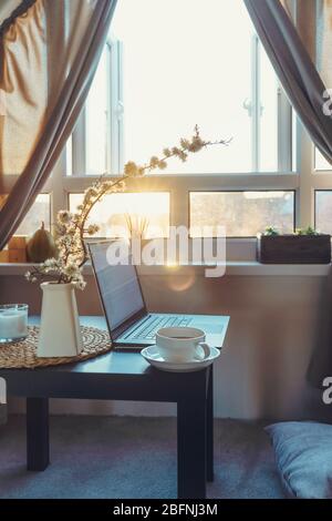Arbeitsplatz mit Laptop, einer Tasse Heißgetränk und einem blühenden Brunch in Vase auf einem Couchtisch in der Nähe des Fensters bei Sonnenuntergang oder Sonnenaufgang. Freiberuflich, arbeitet von ho Stockfoto