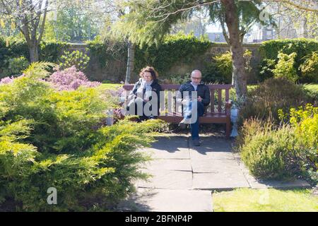 Ein Paar sitzt auf einer Bank in einem leeren Stadtgarten und genießt die Frühlingssonne während der Covid-19-Sperre. Stockfoto