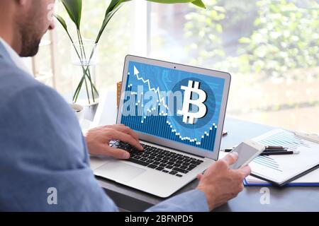 Mann mit Laptop mit Bitcoin-Symbol auf dem Bildschirm und Smartphone am Tisch im Büro Stockfoto