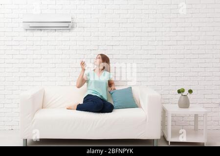 Junge Frau, die die Klimaanlage anschaltet, während sie zu Hause auf dem Sofa sitzt Stockfoto