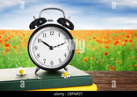 Wecker mit Büchern und Blumen auf Holztisch vor Landschaftshintergrund. Konzept der Zeitänderung Stockfoto