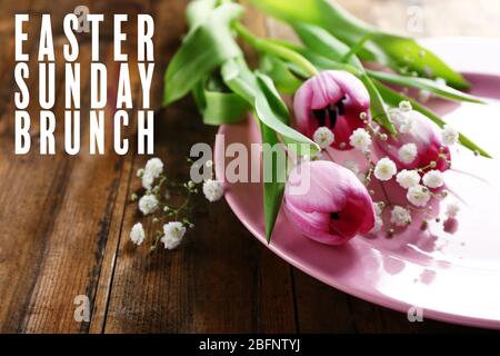 Text OSTERSONNTAG BRUNCH im Hintergrund. Teller und Blumen auf Holztisch Stockfoto