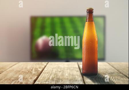 Unbeschriftete Bierflasche auf Holztisch vor der TV-Show von Fußball. Fußballspiel zu Hause ansehen. Stockfoto