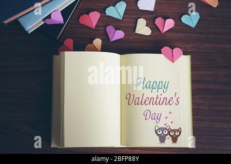 Notizbuch mit Text Happy Valentinstag auf Seite geöffnet. Bunte Origami-Herzen auf Holztisch. Stockfoto
