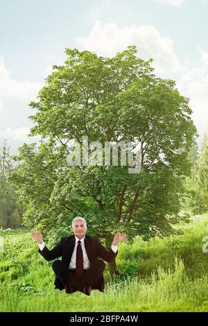Geschäftsmann sitzt in Lotushaltung vor grünem Baumhintergrund. Yoga-Konzept. Stockfoto