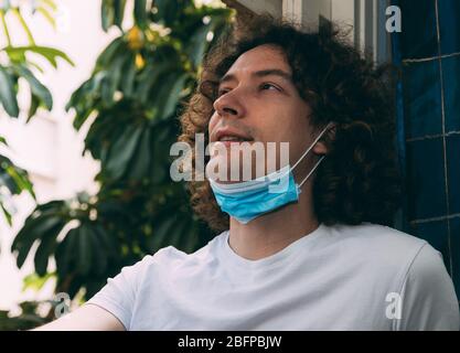 Ein junger Mann mit lockigen langen Haaren, dessen Gesicht eine medizinische Maske abgenommen hat, schaut aus dem Fenster und lächelt. Ende der Heimatquarantäne und des Coronavir Stockfoto