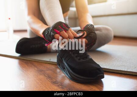 Junge Frau, die während der Quarantäne im Zimmer Sport macht. Nahaufnahme von schwarzen stilvollen Sneakers. Frau bindet Schnürsenkel und bereitet sich auf das Heimtraining vor. Stockfoto