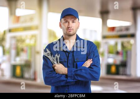 Junger Mechaniker in Uniform mit gekreuzten Armen und Schraubenschlüsseln auf verschwommener Tankstelle Stockfoto