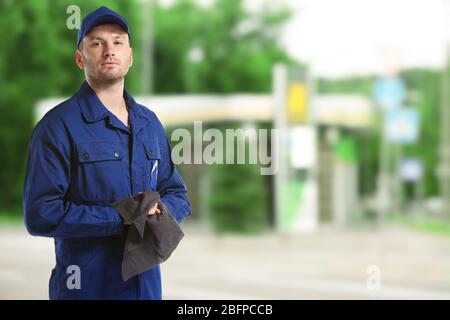 Junger Mechaniker in Uniform mit Lappen auf verschwommener Tankstelle stehend Stockfoto