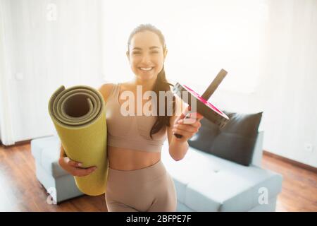 Junge Frau, die während der Quarantäne im Zimmer Yoga-Training macht. Fröhliche positive Mädchen lächelnd und hält gerollte Yoga-Matte und Bauch-Übung Roller Stockfoto