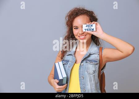 Afroamerikanischer Student hält Bücher und Visitenkarte mit Text IN UNSEREM TEAM auf grauem Hintergrund. Bildungskonzept Stockfoto
