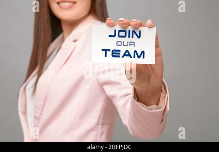 Junge Frau mit Visitenkarte mit Text TRITT UNSEREM TEAM auf grauem Hintergrund bei. Einstellungskonzept Stockfoto