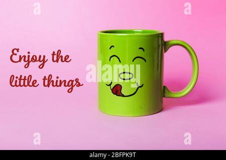 Text GENIESSEN SIE DIE KLEINEN DINGE und Tasse mit lustigen Gesicht auf farbigen Hintergrund Stockfoto