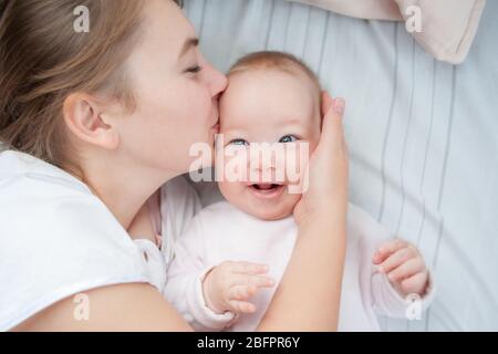Mutter küsst ihr Baby im Bett. Glückliche Mutter küsst Baby auf dem Bett liegen. Schönes lächelndes Mädchen auf dem Rücken liegend. Draufsicht. Flaches Lay Stockfoto