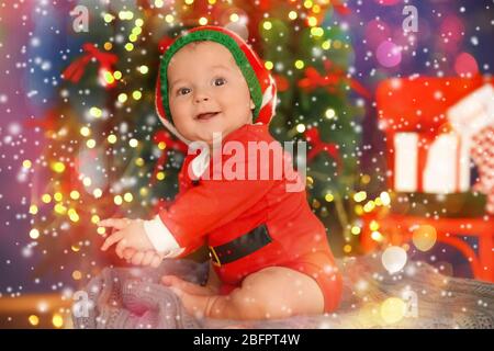 Niedliches Baby im Kostüm des Weihnachtshelfers und Weihnachtsbaum auf Hintergrund. Festtage Konzept Stockfoto