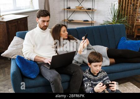 Mutter, Vater und Sohn mit Laptop und Handy zu Hause. Familienmitglieder ignorieren einander und sprechen live. Gadget Einfluss auf Familie Stockfoto