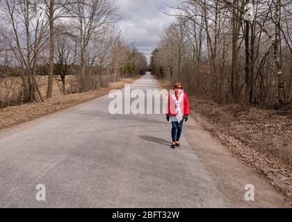 Eine Frau, die einen leuchtend roten Mantel trägt, geht allein eine Landstraße während der Covid-19-Pandemie entlang. Stockfoto