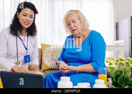 Lächelnde junge Ärztin, die ältere Patienten zu Hause besucht und die Ergebnisse medizinischer Tests auf dem Bildschirm des Tablet-Computers zeigt Stockfoto